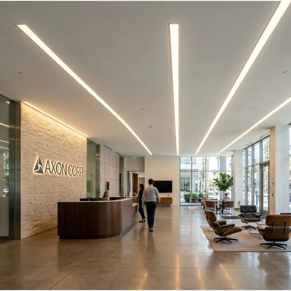 シームレスな4インチ幅の埋込線形LED照明が設置されたホワイトの石膏天井を持つ高級クラスAオフィスロビーの広角ビューです。長く連続した光のラインが木製の受付デスクや石のアクセントウォールへと視線を誘導し、プロフェッショナルで歓迎的な商業空間を演出します。.