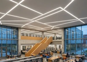 A spacious modern university library with a high drywall ceiling featuring 4-inch recessed linear lights arranged in a creative, intersecting geometric pattern. The lighting provides functional task light for study areas and bookshelves while serving as a bold architectural design element.